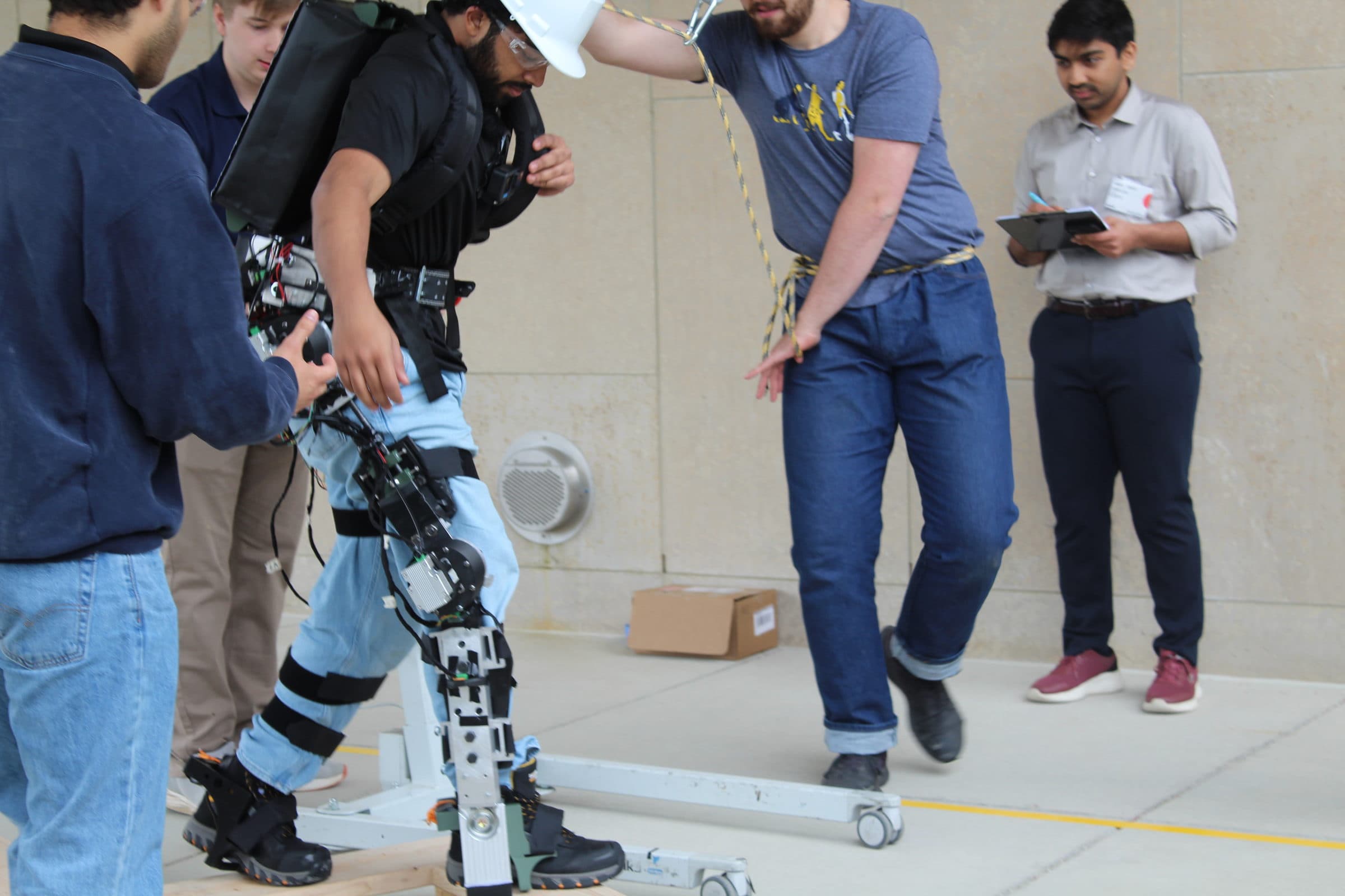 A pilot in the McMaster Exoskeleton suit being supported by team members during an obstacle course run.